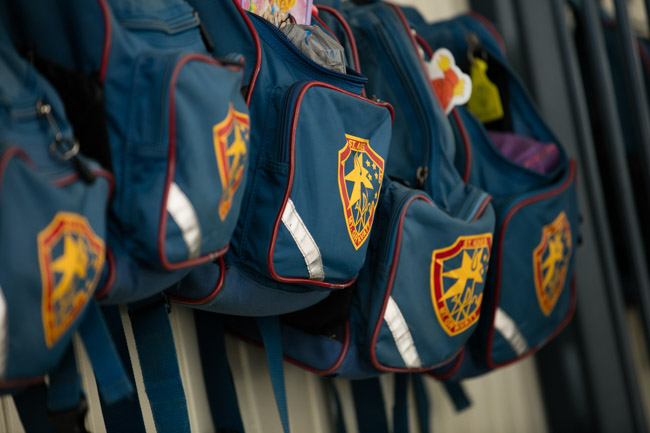 Row of blue school backpacks with school crests hanging on hooks along a wall.