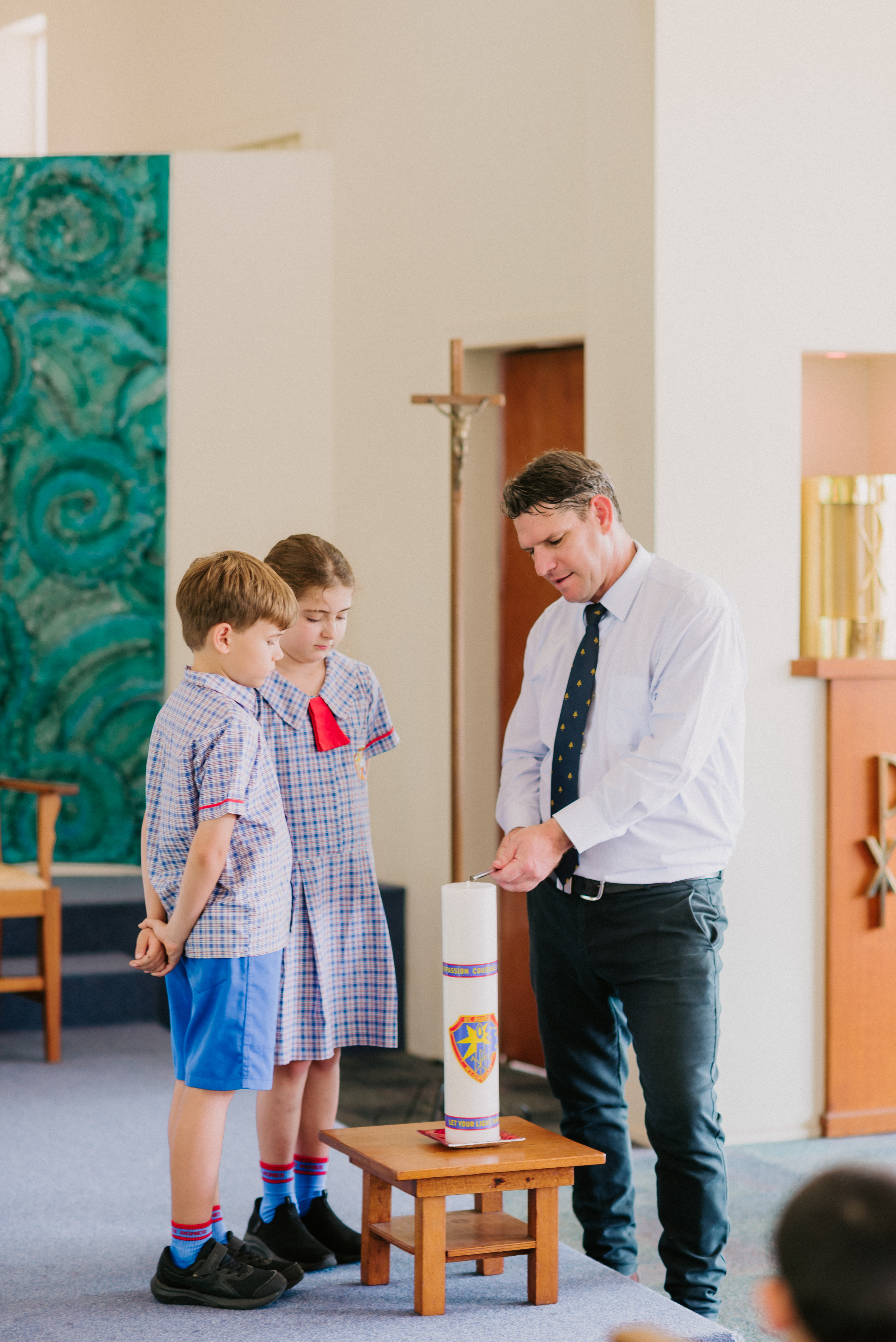 Adult lighting a candle on a stand while two students in school uniform stand beside it during a school liturgy inside a church.