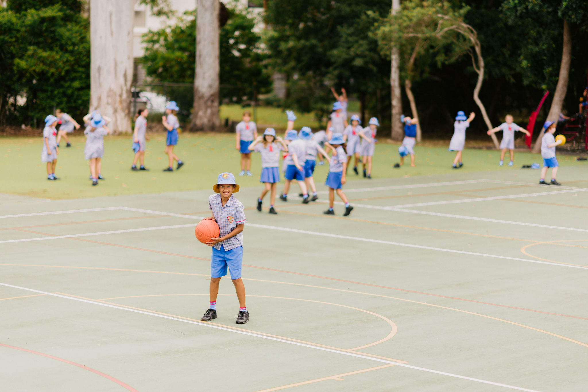 Student holding a basketball on an outdoor school court, with other students participating in a group activity in the background.