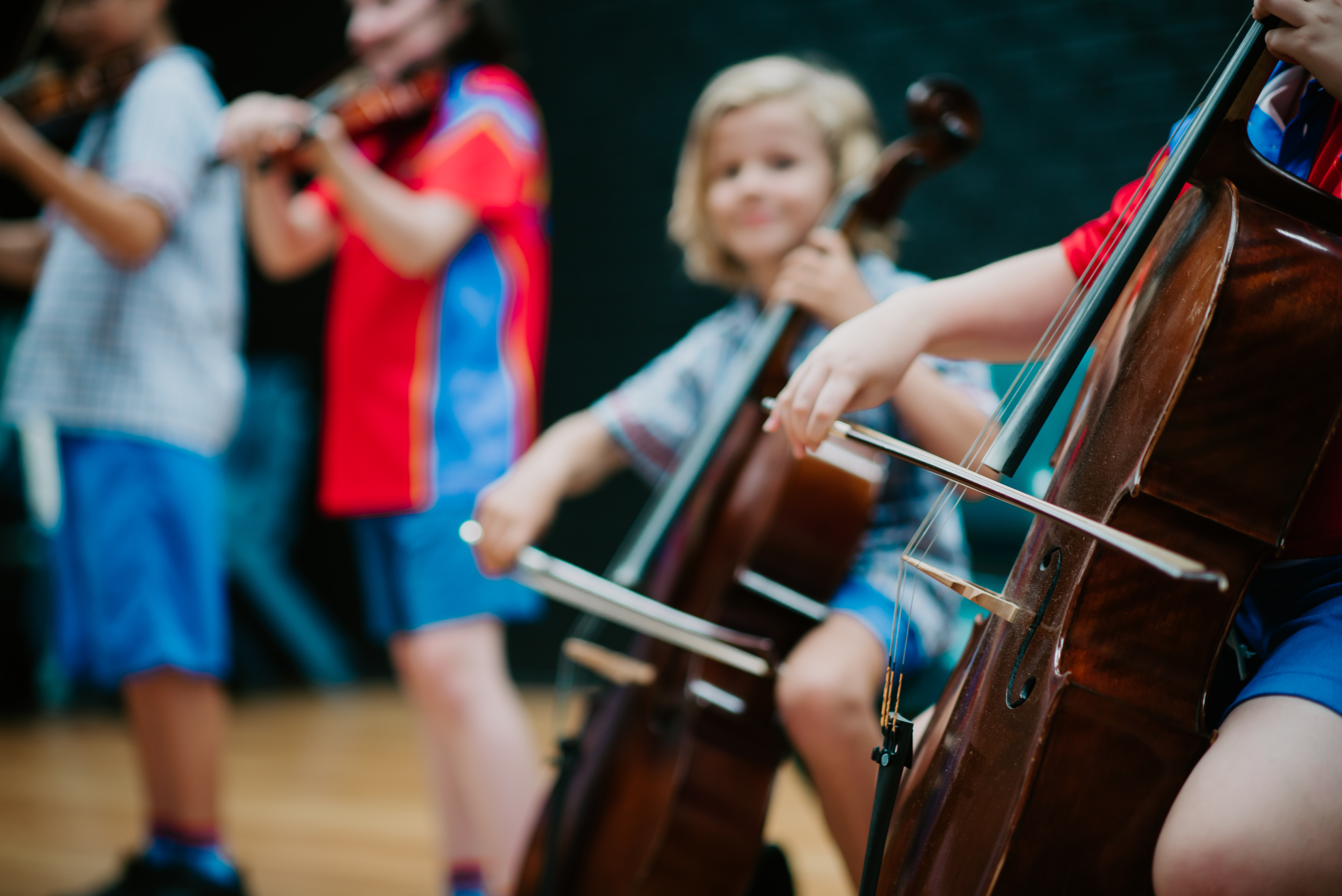 Close-up of a student bowing a cello while other students play violins in the background during a school music lesson.