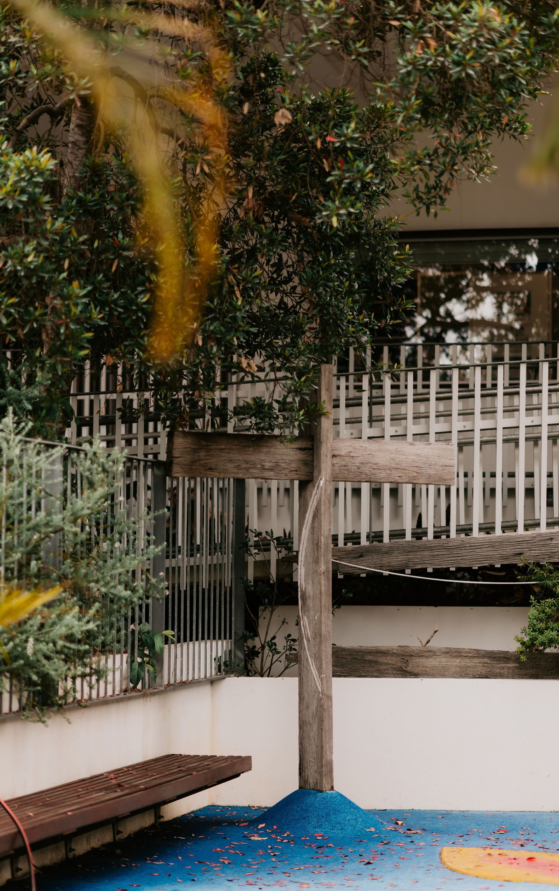 Outdoor school courtyard with a wooden bench, railing, and shaded walkway surrounded by trees and shrubs.