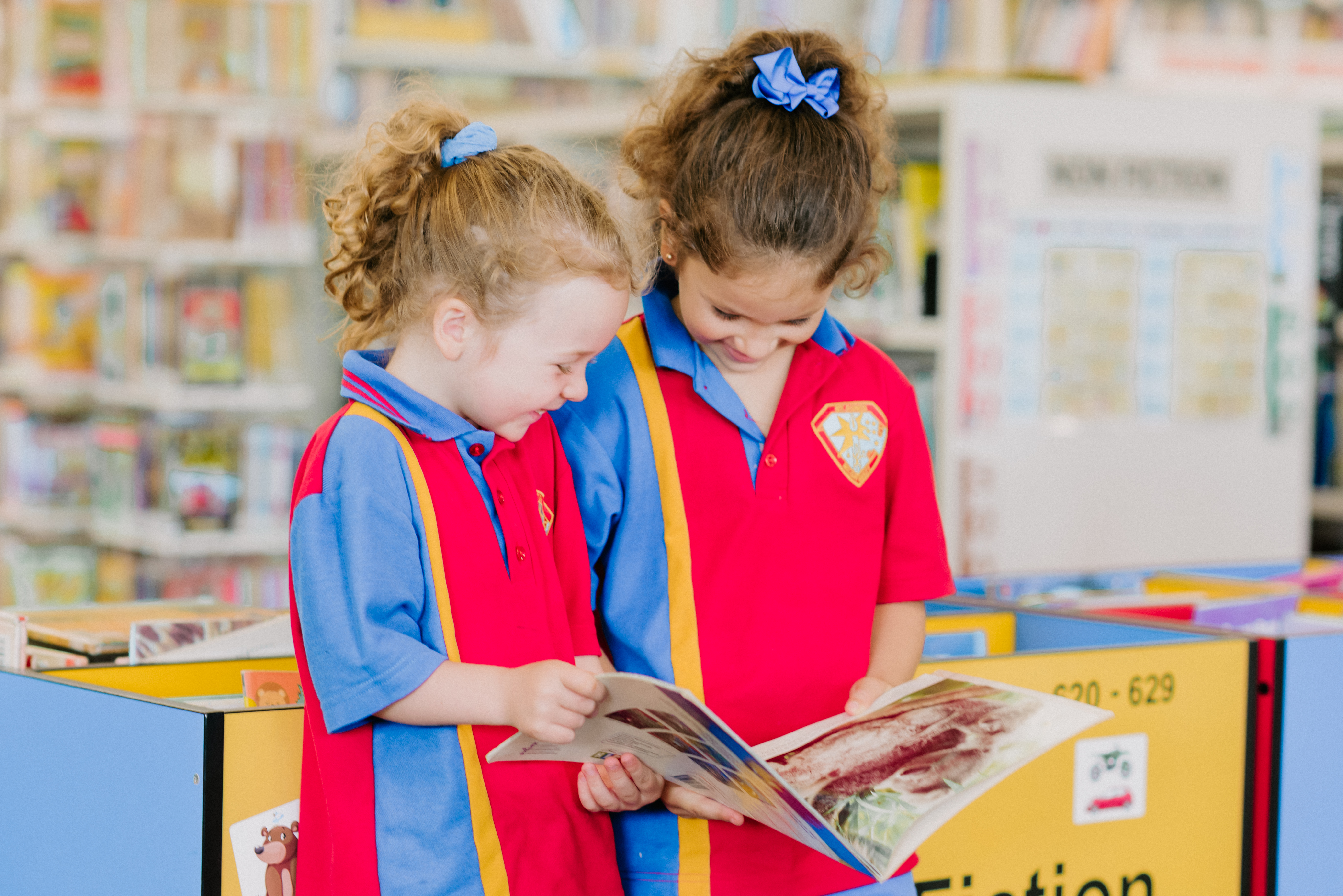 Two young students in school uniform standing together in a library and reading a book.