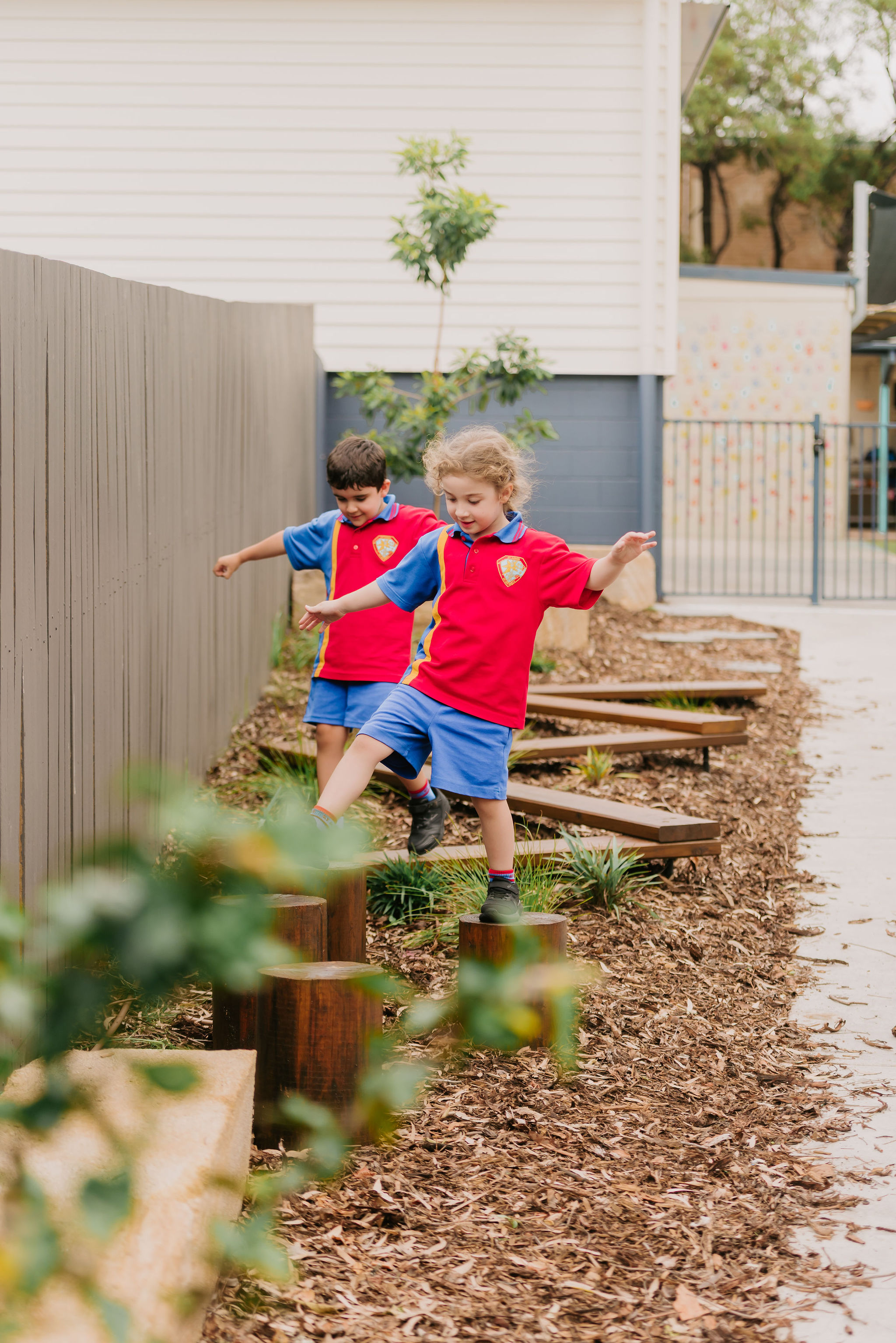 Two students in school uniform balancing on wooden stepping posts in an outdoor nature‑play area.