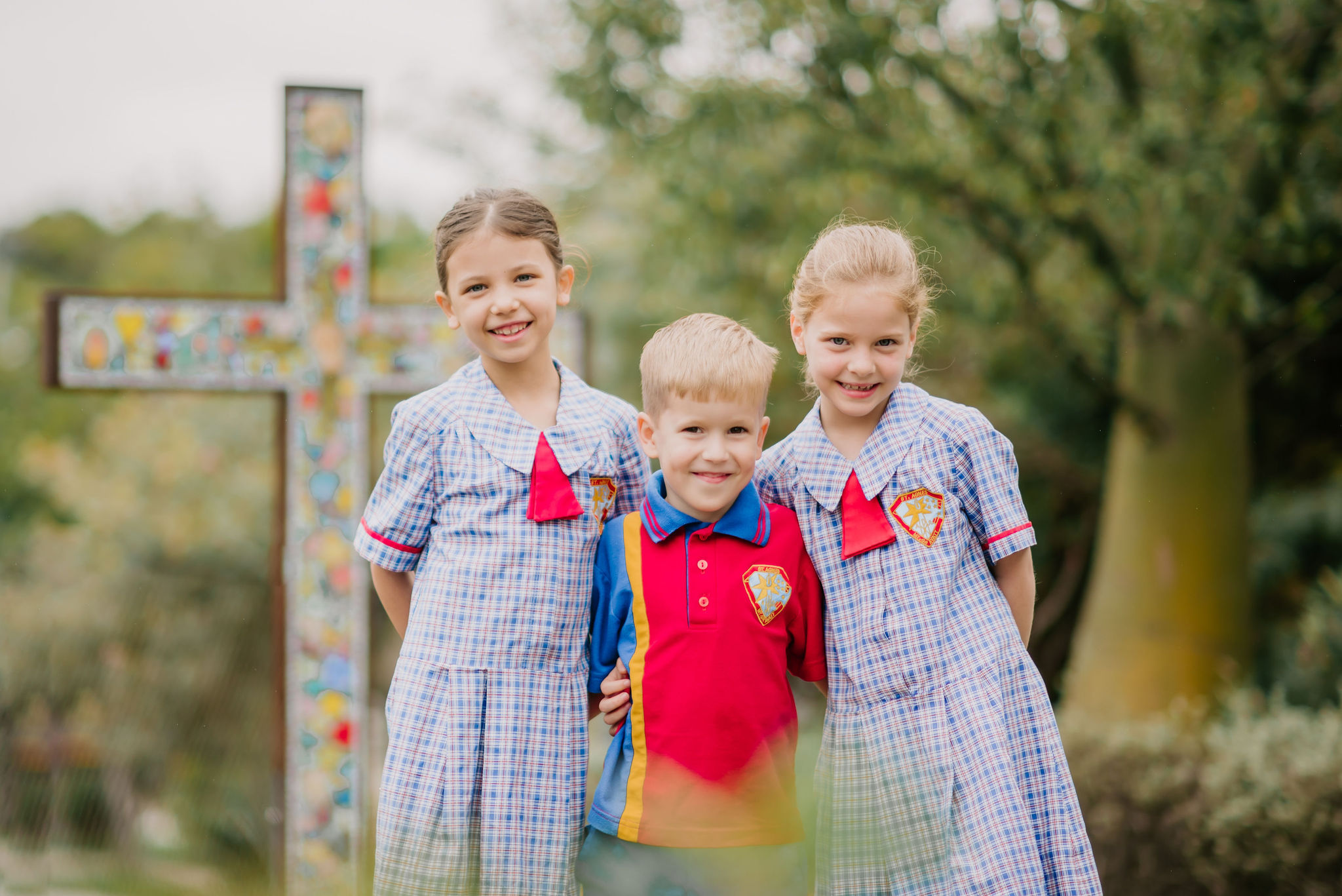 Three students in school uniform standing together outdoors in front of a large decorative cross.