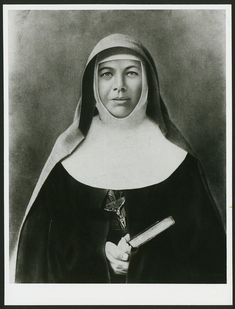 Historic black‑and‑white portrait of a nun in religious habit holding a book, with a crucifix visible at the chest.