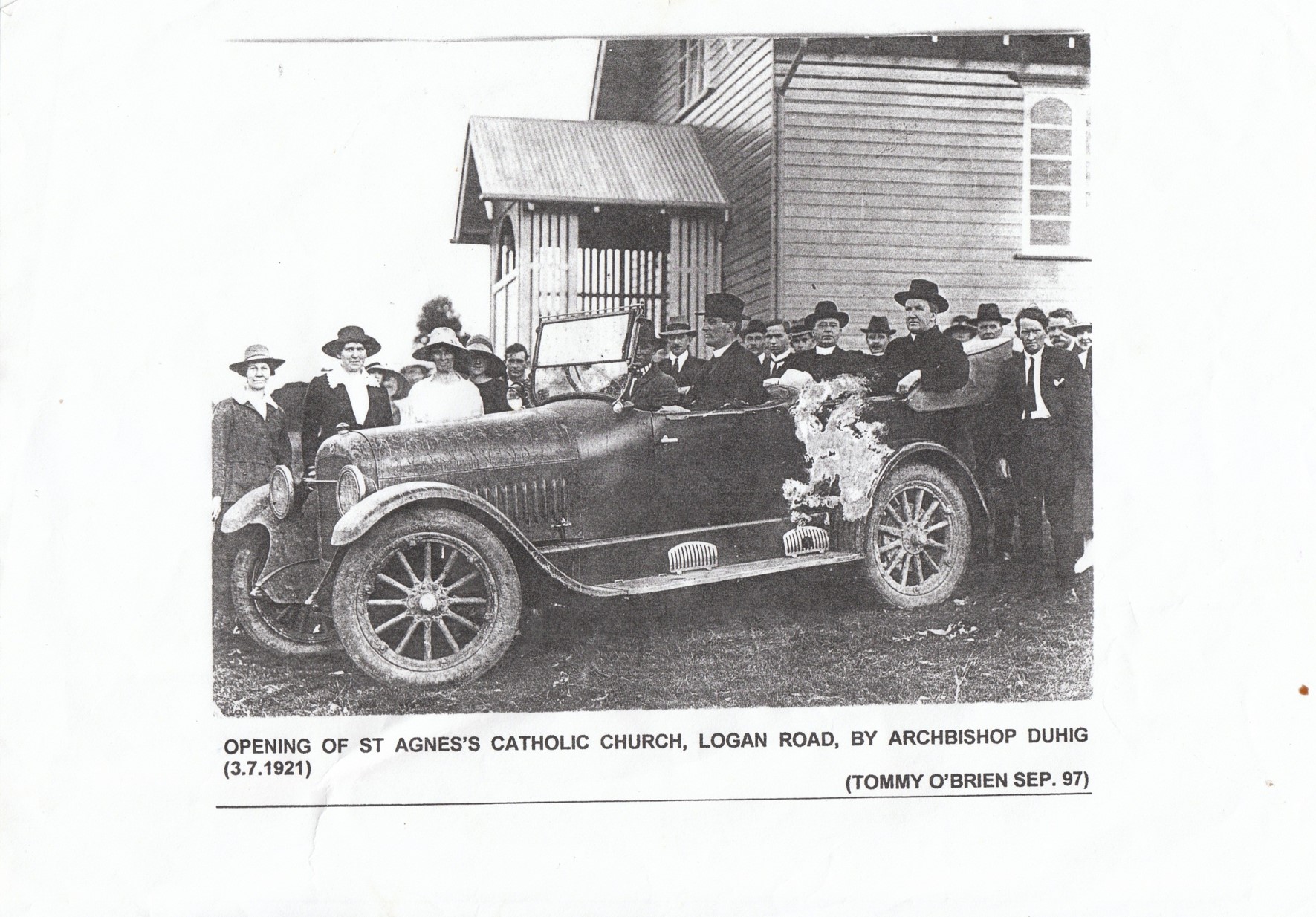Historic black‑and‑white photograph showing a crowd gathered around an early motor car outside a timber church building.