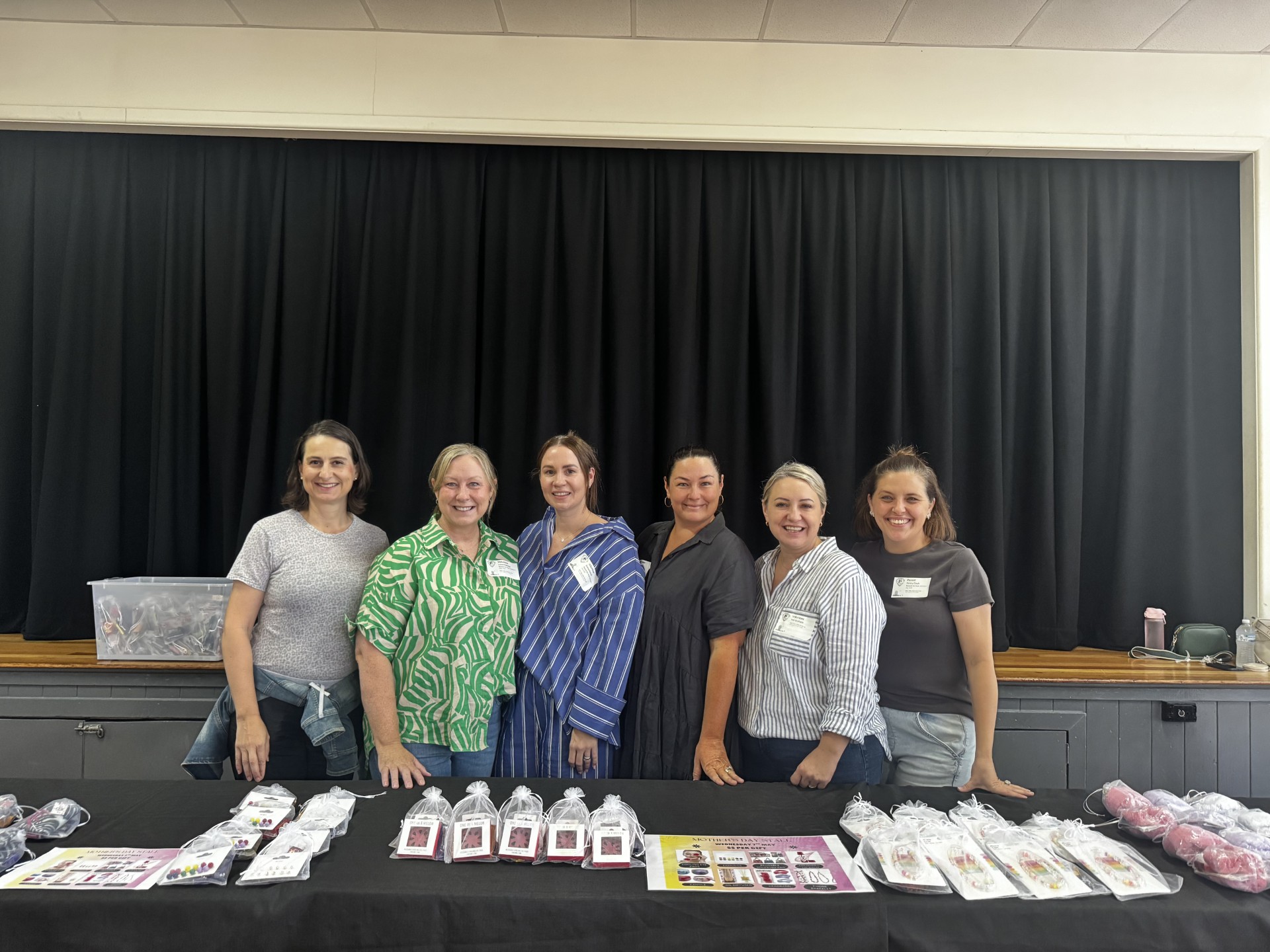 School staff standing behind a display table with handmade items prepared for a school event inside a hall.