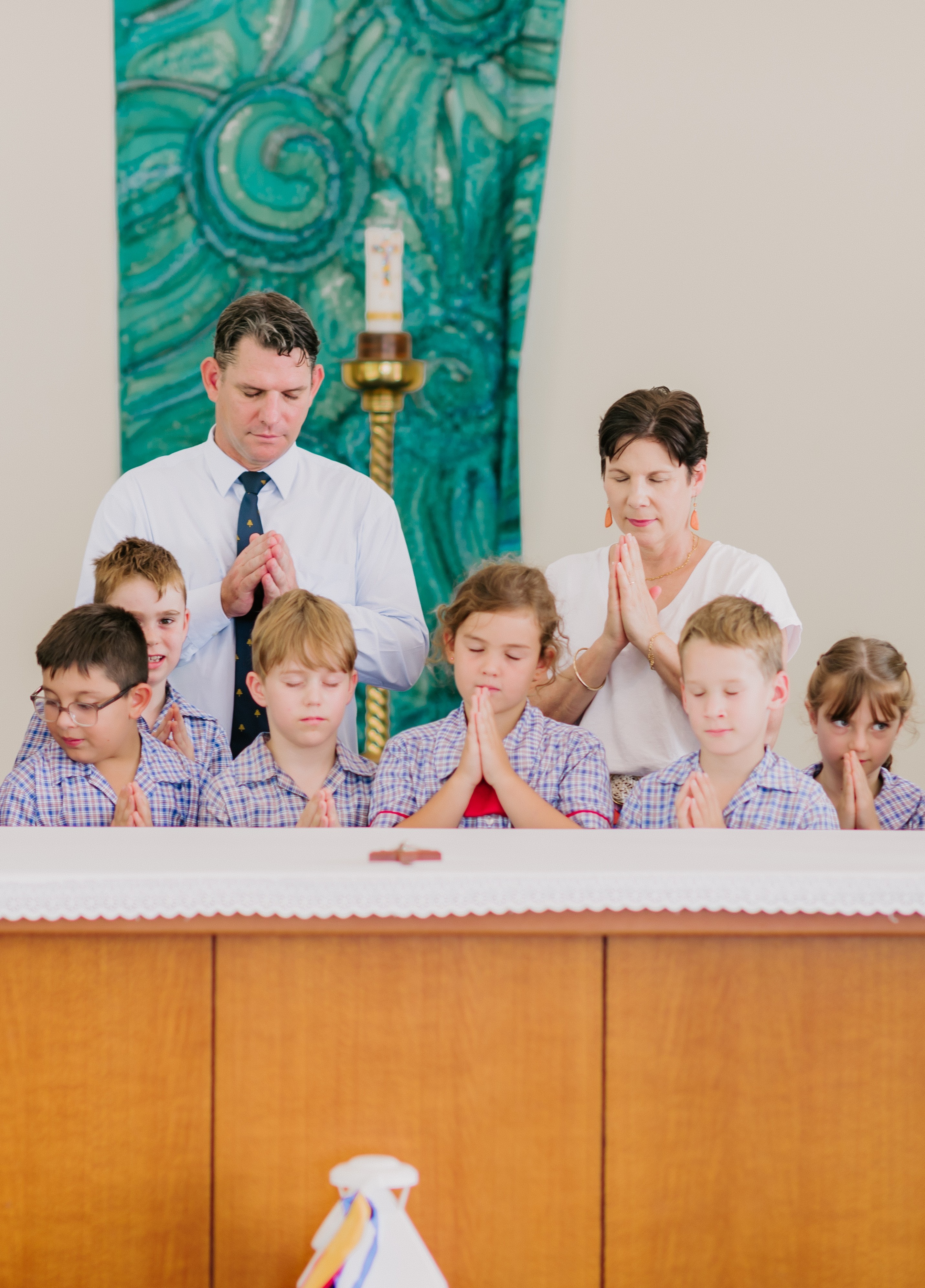 Students in school uniform standing with adults at the front of a church with hands together during a prayer service.
