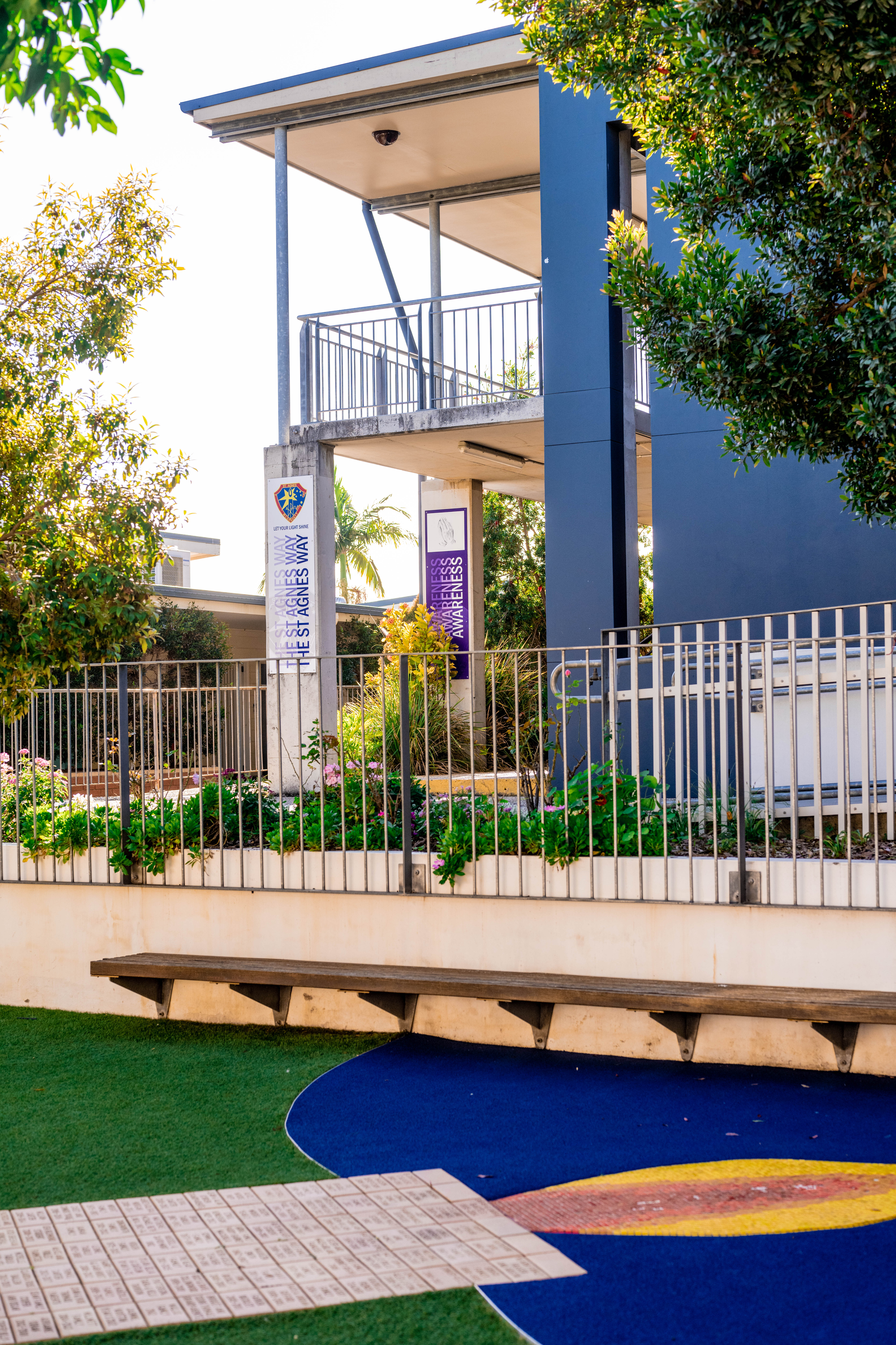 Modern school building with an upper balcony, landscaped garden beds, and playground surfaces with coloured markings in the foreground.