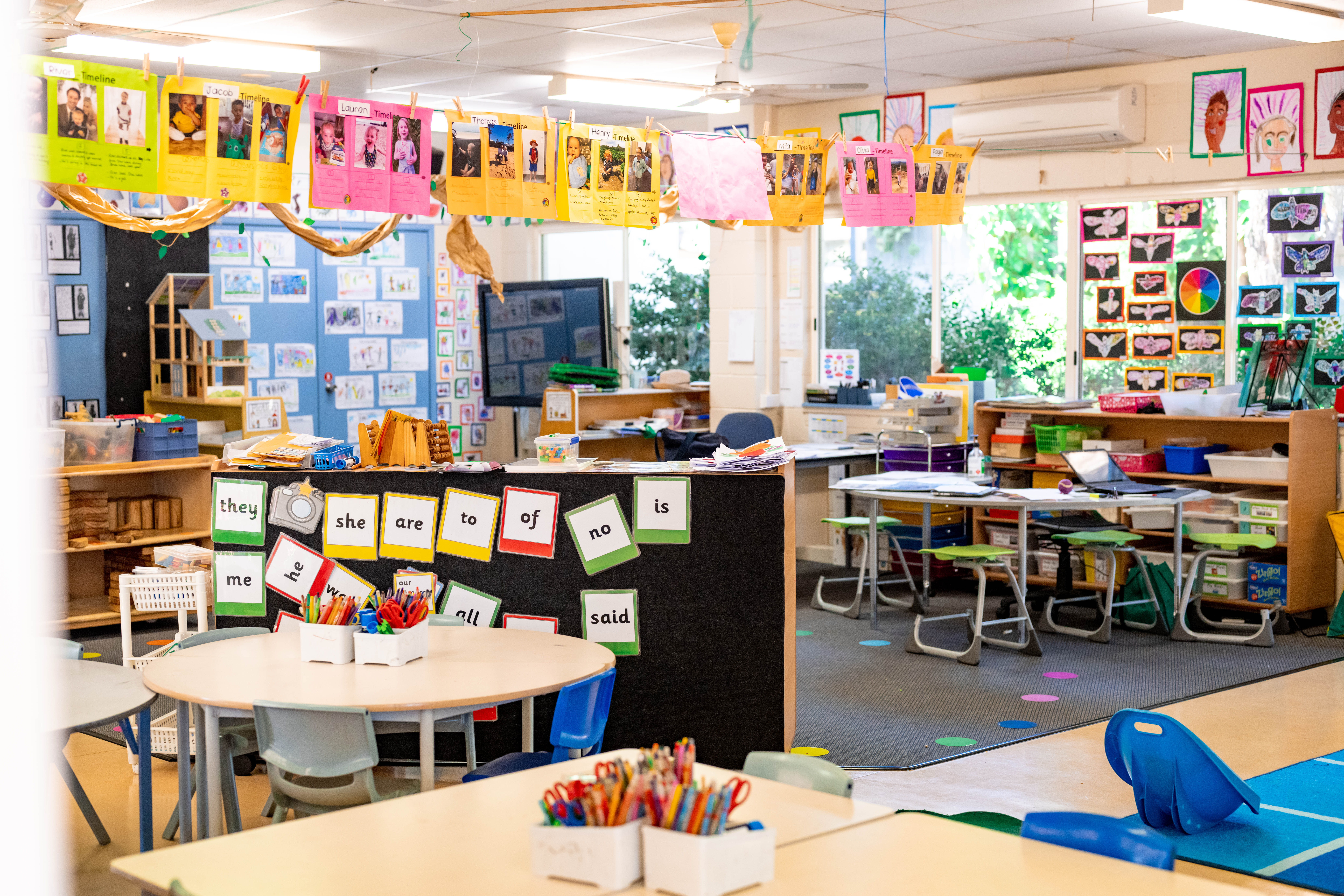 Bright classroom with tables and chairs, student artwork displayed on walls and hanging lines, and word cards arranged on a central partition.
