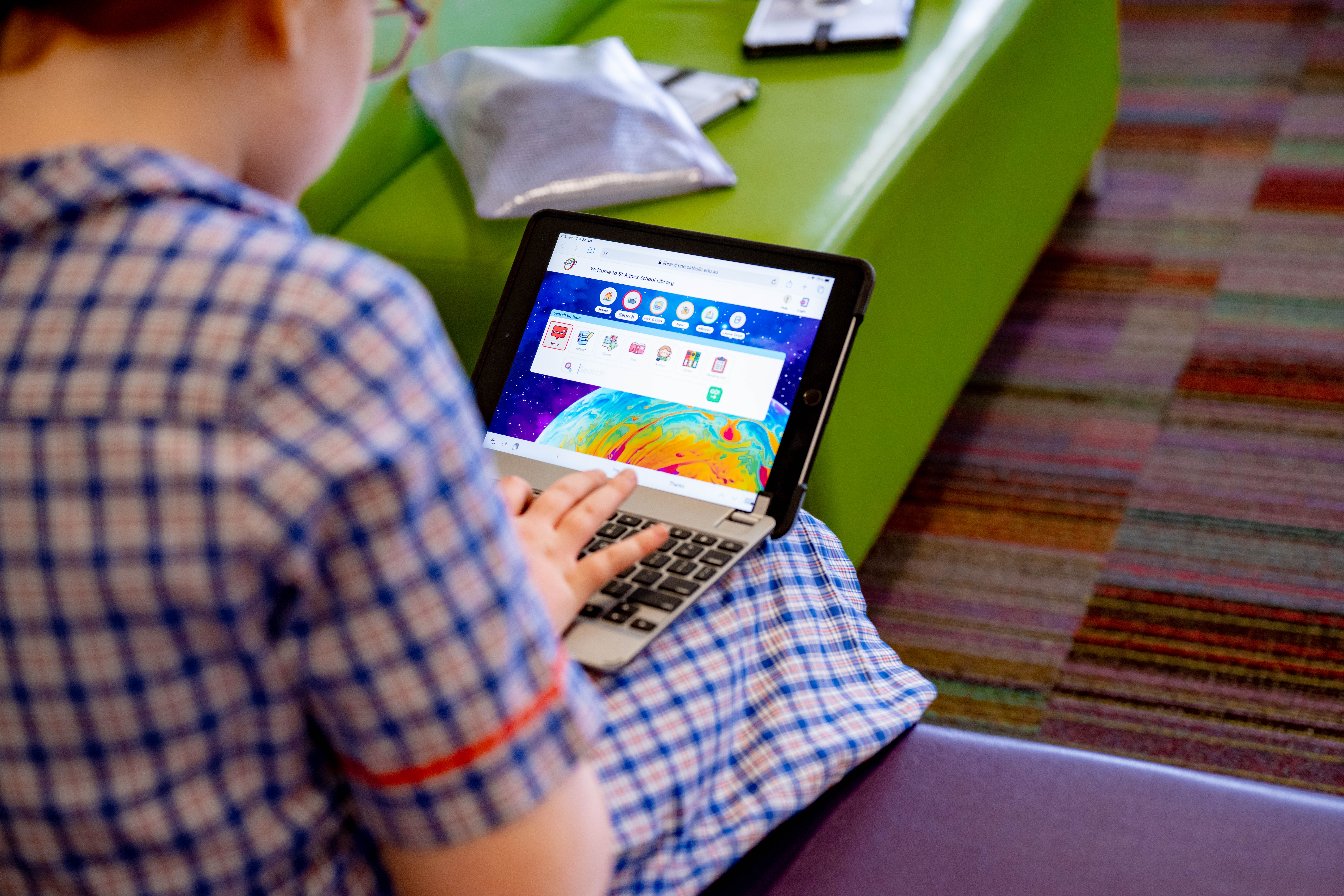 Student in school uniform using a tablet with a keyboard case while seated in a school learning space.