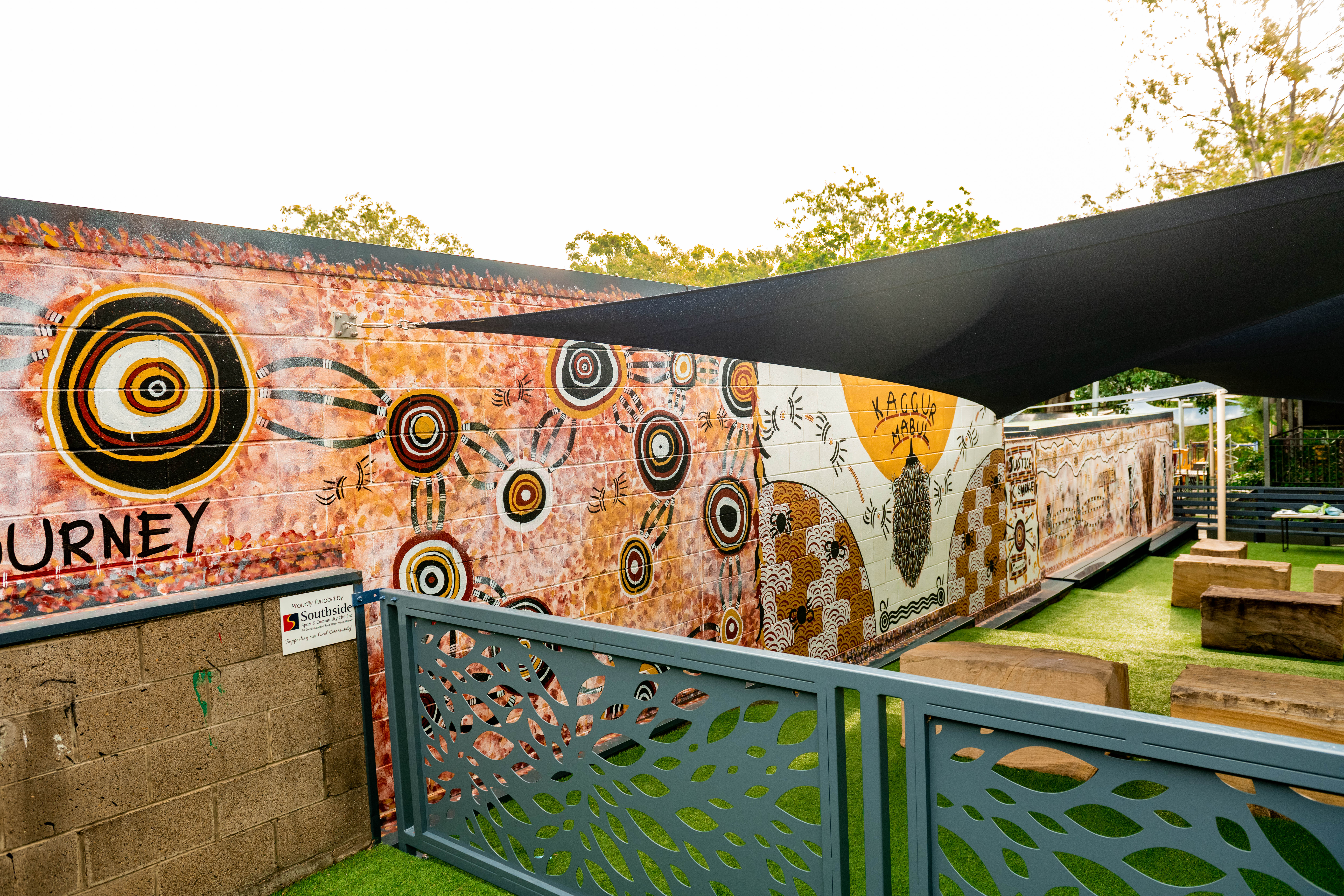 Outdoor learning area with a wall mural featuring circular patterns and symbols, shaded seating, and a fenced play space.